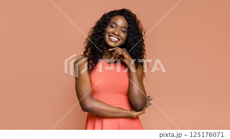 This image shows a Black woman with curly hair, wearing a coral sleeveless dress and smiling, against a peach-colored background. She has her arms crossed and is looking directly at the camera. 125176071