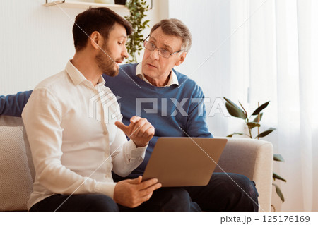 Adult Son Teaching Senior Father To Use Laptop Explaining How To Browse Internet Sitting Together On Couch Indoor. Selective Focus 125176169
