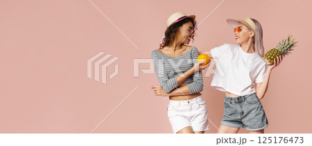 This image shows two friends in casual summer clothing, standing in front of a pink wall and smiling at each other. One woman is holding an orange, and the other is holding a pineapple This image shows two friends in casual summer clothing, standing in front of a pink wall and smiling at each other. One woman is holding an orange, and the other is holding a pineapple 125176473