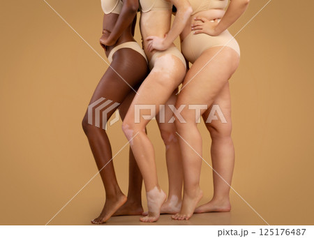 Body positivity. Three ladies with owerweight, vitiligo and stretch marks posing together in underwear, standing in lingerie over beige background in studio, side view, cropped image with copy space Body positivity. Three ladies with owerweight, vitiligo and stretch marks posing together in underwear, standing in lingerie over beige background in studio, side view, cropped image with copy space 125176487