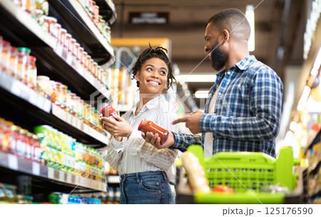 Happy African Family Couple Buying Food In Supermarket, Choosing Products Walking With Cart Along Aisles And Full Shelves Purchasing Groceries Together. Black Spouses Purchasing Essentials Together 125176590