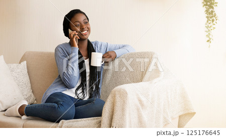 Smiling African American Woman Talking on Cellphone And Drinking Morning Coffee While Sitting On Couch At Home 125176645