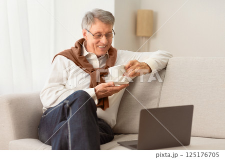 Retirement Lifestyle. Happy Elderly Man Using Laptop Watching Movie Online And Having Coffee Sitting On Couch At Home. Selective Focus Retirement Lifestyle. Happy Elderly Man Using Laptop Watching Movie Online And Having Coffee Sitting On Couch At Home. Selective Focus 125176705