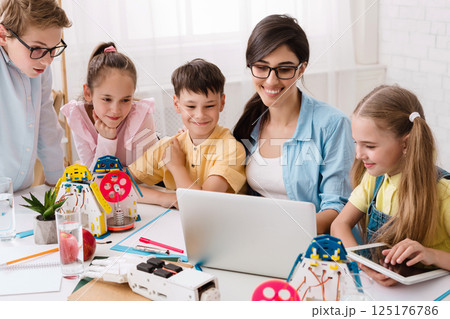 Elementary school students are working on a robotics project in their classroom. The students are gathered around a laptop and are looking at the screen intently. Their teacher is sitting beside them 125176786