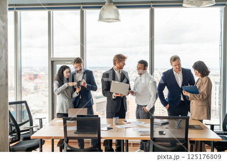 A group of business professionals stand around a large table in a modern office. There is a view of a city through the large windows behind them. A group of business professionals stand around a large table in a modern office. There is a view of a city through the large windows behind them. 125176809
