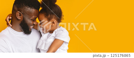 Black father and his daughter share a tender moment, smiling and laughing while embracing each other. The warm yellow backdrop highlights their joyful expressions and creates a cheerful atmosphere 125176865