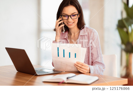 Checking Papers. Young businesswoman in glasses talking on mobile phone, looking at document with graphs, copyspace Checking Papers. Young businesswoman in glasses talking on mobile phone, looking at document with graphs, copyspace 125176866