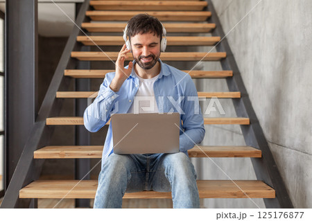 A man sits on a staircase with a laptop in his lap. He is wearing headphones and adjusting them while looking at the screen. He is wearing a blue button down shirt and jeans. A man sits on a staircase with a laptop in his lap. He is wearing headphones and adjusting them while looking at the screen. He is wearing a blue button down shirt and jeans. 125176877