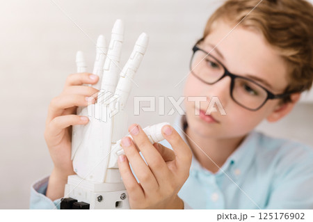 A young boy wearing glasses and a light blue shirt examines a white robotic hand. He is holding the robotic hand with both hands and looking at it intently. A young boy wearing glasses and a light blue shirt examines a white robotic hand. He is holding the robotic hand with both hands and looking at it intently. 125176902