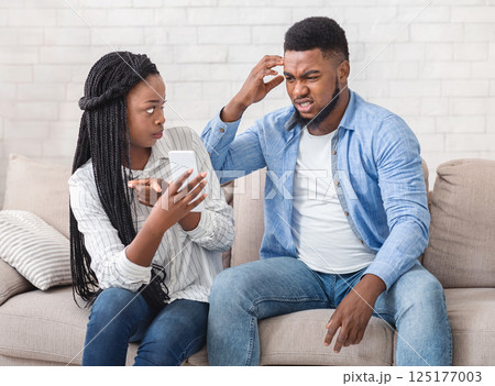 Infidelity Concept. Angry Black Girlfriend Showing Her Cheating Boyfriend His Cellphone, Demanding Explanation Sitting On Couch At Home. 125177003