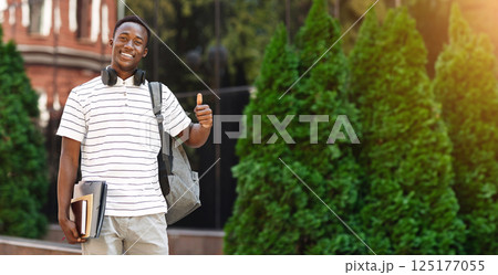 Portrait of happy black male student with backpack and workbooks posing outdoors and showing thumb up gesture, copy space Portrait of happy black male student with backpack and workbooks posing outdoors and showing thumb up gesture, copy space 125177055