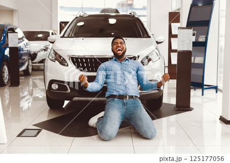 New Auto. Excited African Man Shouting Shaking Fists Celebrating Buying Car Standing On Knees In Dealership Store New Auto. Excited African Man Shouting Shaking Fists Celebrating Buying Car Standing On Knees In Dealership Store 125177056