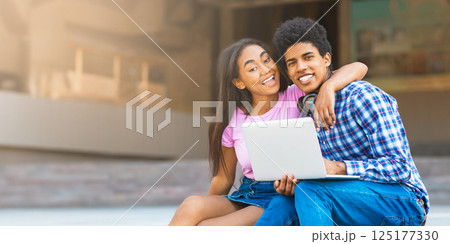 A teen black couple sits together on a ledge in the city, laughing and using a laptop. The girl is wearing a pink shirt and denim skirt, while guy wears a plaid shirt and jeans. A teen black couple sits together on a ledge in the city, laughing and using a laptop. The girl is wearing a pink shirt and denim skirt, while guy wears a plaid shirt and jeans. 125177330