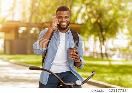Coffee On The Go. Portrait of smiling african american man on bicycle talking on smartphone with cup of hot drink in hands Coffee On The Go. Portrait of smiling african american man on bicycle talking on smartphone with cup of hot drink in hands 125177384