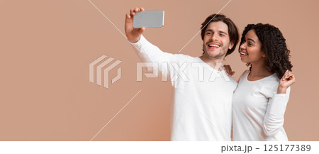 Say Cheese. Cheerful interracial couple taking sellfie on smartphone together, posing over yellow background with free space 125177389