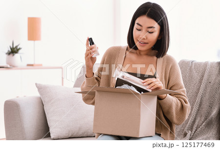 Online Shopping. Smiling asian woman opening parcel box with cosmetics at home, holding makeup products, unpacking gift sitting on the couch at home. Lady satisfied with delivery service Online Shopping. Smiling asian woman opening parcel box with cosmetics at home, holding makeup products, unpacking gift sitting on the couch at home. Lady satisfied with delivery service 125177469