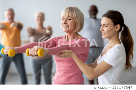 Young lady coach helping senior woman while exercising with dumbbells, sporty multiracial group of elderly people having fitness class at nursing home, training with instructor Young lady coach helping senior woman while exercising with dumbbells, sporty multiracial group of elderly people having fitness class at nursing home, training with instructor 125177544