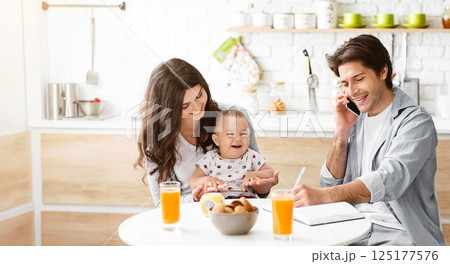A happy family enjoys a casual breakfast together in their kitchen. A young woman holds her baby in her lap, and a man sits at the table, smiling while talking on the phone. A happy family enjoys a casual breakfast together in their kitchen. A young woman holds her baby in her lap, and a man sits at the table, smiling while talking on the phone. 125177576