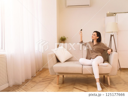 Split System. Satisfied european girl holding remote control relaxing under the air conditioner, sitting on couch Split System. Satisfied european girl holding remote control relaxing under the air conditioner, sitting on couch 125177666