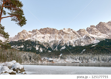 Picturesque Alpine Ski Resort Nestled between Frozen Lake and Snow Covered Mountain Peaks with Traditional Wooden Chalets Reflecting in Crystal Clear Water Surface during Winter Season in European 125177755