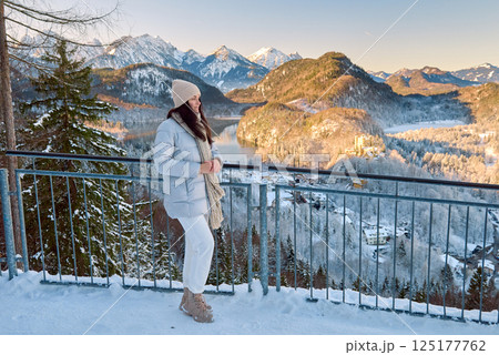Beautiful young woman in a white winter coat standing on a balcony overlooking snow-covered mountains near Neuschwanstein Castle in Bavaria, Germany 125177762