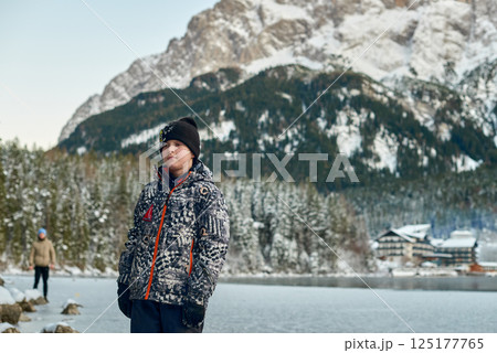 Young boy standing on a frozen lake in a snowy winter landscape with evergreen forest and majestic mountains in the background on a clear cold day 125177765