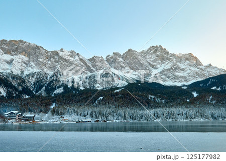 Picturesque Alpine Ski Resort Nestled between Frozen Lake and Snow Covered Mountain Peaks with Traditional Wooden Chalets Reflecting in Crystal Clear Water Surface during Winter Season in European 125177982