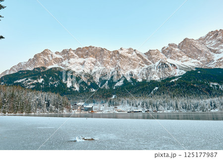 Picturesque Alpine Ski Resort Nestled between Frozen Lake and Snow Covered Mountain Peaks with Traditional Wooden Chalets Reflecting in Crystal Clear Water Surface during Winter Season in European 125177987