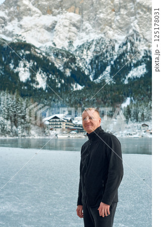 Middle-aged man in black jacket standing in serene snowy mountain landscape with snow-capped peaks and evergreen forests, reflecting tranquil winter atmosphere. 125178031
