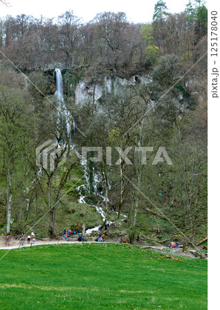 Tranquil Green Meadow with Winding Path to Cascading Waterfall, Rocky Cliff, Bare Trees, Overcast Sky, Serene Atmosphere. 125178040