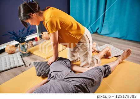 Masseuse making thai yoga massage. Woman therapist doing traditional massage treatment, stretching male patient's body. Client lying on mat, enjoying hand massage in calm atmosphere. Masseuse making thai yoga massage. Woman therapist doing traditional massage treatment, stretching male patient's body. Client lying on mat, enjoying hand massage in calm atmosphere. 125178385