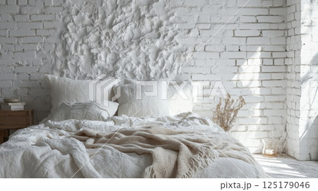 A white bed with white linens and a beige throw blanket is positioned against a white brick wall. Sunlight streams in through a window, illuminating the room 125179046