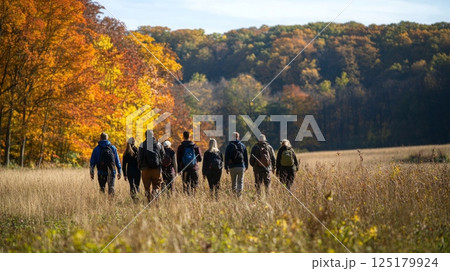 A diverse group of enthusiasts walks together along a grassy path, surrounded by colorful autumn foliage under a clear blue sky. The warm sunlight enhances the fall colors. 125179924