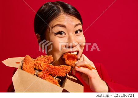 Close-up of emotional Asian woman biting spicy crispy chicken nuggets with tomato sauce from carton box to go against red background. 125180721