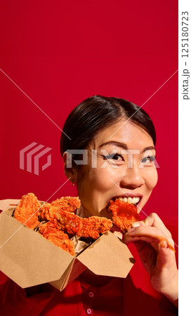 Food delivery. Close-up of emotional Asian woman eating delicious crispy chicken nuggets with tomato sauce from carton box against red background. 125180723