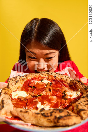 Woman in red bites giant pizza slice with intense hunger and playful face against bright yellow background. Delicious Italian cuisine. Woman in red bites giant pizza slice with intense hunger and playful face against bright yellow background. Delicious Italian cuisine. 125180826