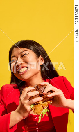 Portrait of young Asian woman in red emotionally biting delicious burger with sauce of her mouth against bright yellow background. Portrait of young Asian woman in red emotionally biting delicious burger with sauce of her mouth against bright yellow background. 125180871