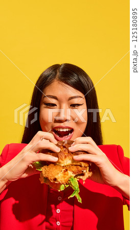Excited Asian young woman in red prepares to bite large sandwich while looking at it with open mouth and wide smile against bright yellow background. 125180895