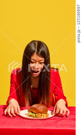Young woman in red outfit leans over burger with hungry gaze on red table against yellow background. Emotional addiction to food Young woman in red outfit leans over burger with hungry gaze on red table against yellow background. Emotional addiction to food 125180897