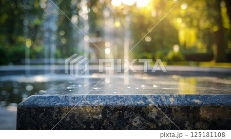 A close-up of a granite fountain edge with water droplets and a blurred background of green trees and a fountain in the distance. 125181108