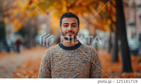 A young man stands confidently in a cozy sweater, surrounded by vibrant autumn leaves in a city park. The scene captures the essence of a pleasant day, with warm colors and a calm atmosphere. 125181719