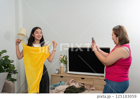 Fashion and Self-Expression. A woman capturing her friend in a vibrant yellow dress, showcasing their style in a cozy home setting. 125183059