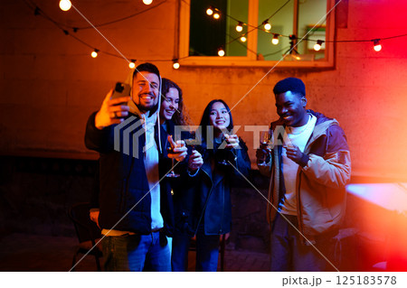Friends enjoying a joyful gathering outdoors at night with drinks under string lights Friends enjoying a joyful gathering outdoors at night with drinks under string lights 125183578