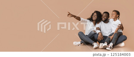 Happy black family of three sitting on floor and pointing aside at copy space over yellow background, panorama Happy black family of three sitting on floor and pointing aside at copy space over yellow background, panorama 125184003