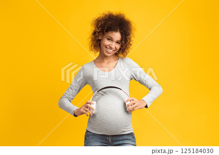 Pregnancy therapy. Afro woman holding headphones on her belly, pink background Pregnancy therapy. Afro woman holding headphones on her belly, pink background 125184030