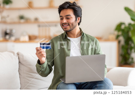 A young Arab man sits on a white couch in his home, holding a credit card in his left hand while looking at a laptop in his right. He smiles, presumably satisfied with his online purchase. A young Arab man sits on a white couch in his home, holding a credit card in his left hand while looking at a laptop in his right. He smiles, presumably satisfied with his online purchase. 125184053