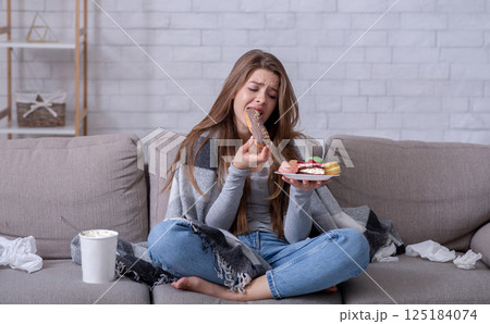 Comfort foods and stress eating concept. Frustrated young lady devouring pastry on sofa at home. Upset woman dealing with negative emotions, anxiety or problems through sweets Comfort foods and stress eating concept. Frustrated young lady devouring pastry on sofa at home. Upset woman dealing with negative emotions, anxiety or problems through sweets 125184074