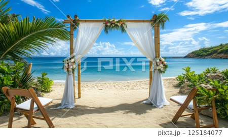 A picturesque wedding altar is set on the sandy beach, adorned with flowers and draped fabric, overlooking calm turquoise waters and a bright blue sky. 125184077