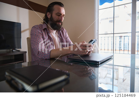 Bearded Man Typing on Phone at Home Office Desk Near Large Window Bearded Man Typing on Phone at Home Office Desk Near Large Window 125184469