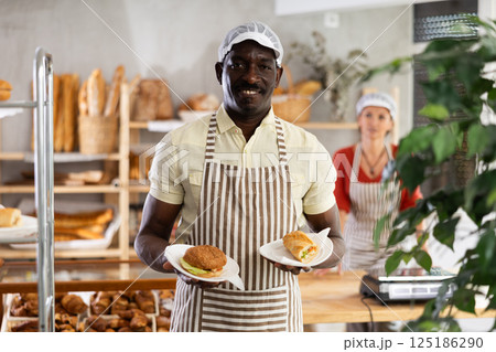 African man entrepreneur, owner of bakery, holds out sandwich in disposable dish plate in hand 125186290